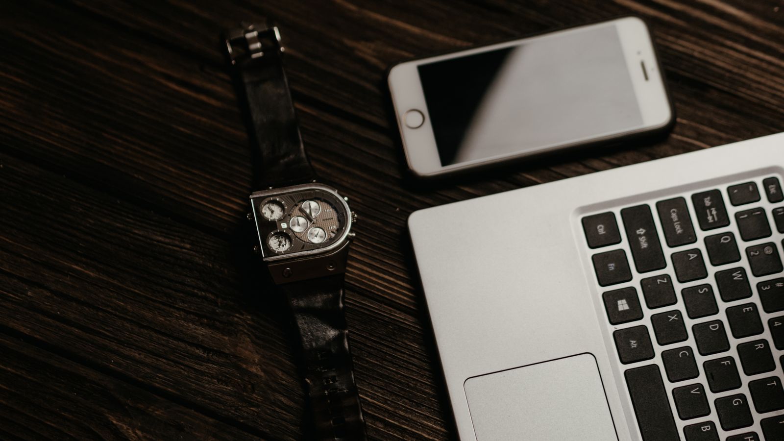 A laptop, smartphone and wristwatch arranged on a wooden table