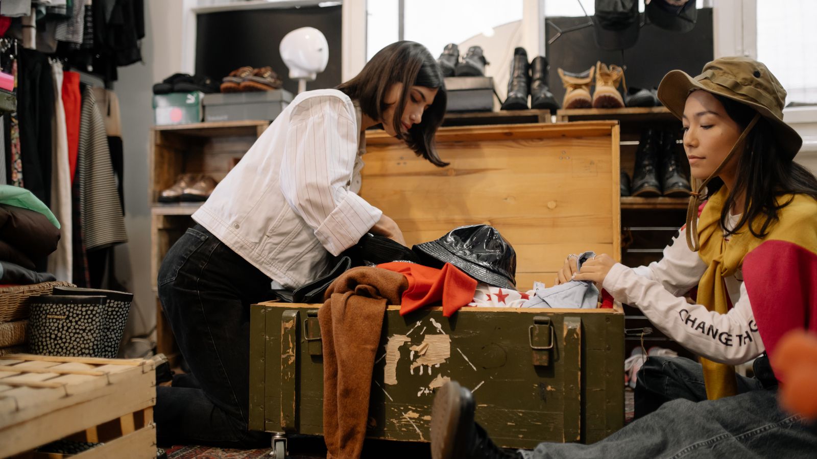 Two women sorting through vintage clothing in a thrift shop full of shoes and hats