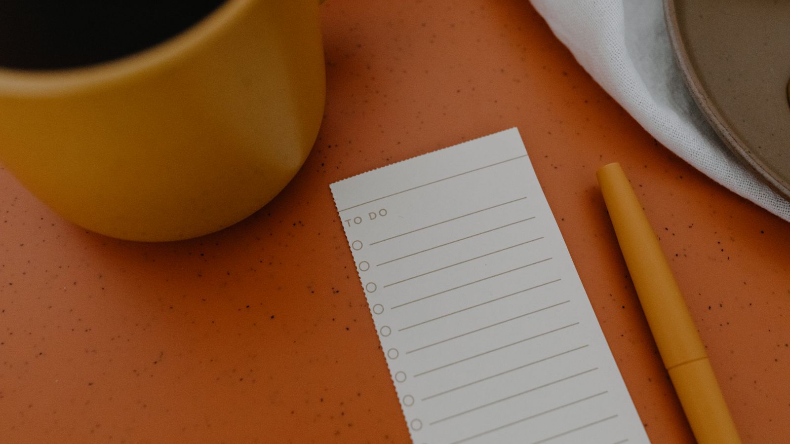 A flat lay of a mug, to-do list and pen on an orange surface