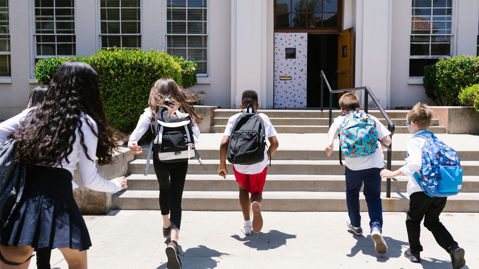 Five children with backpacks running up the front steps of a school