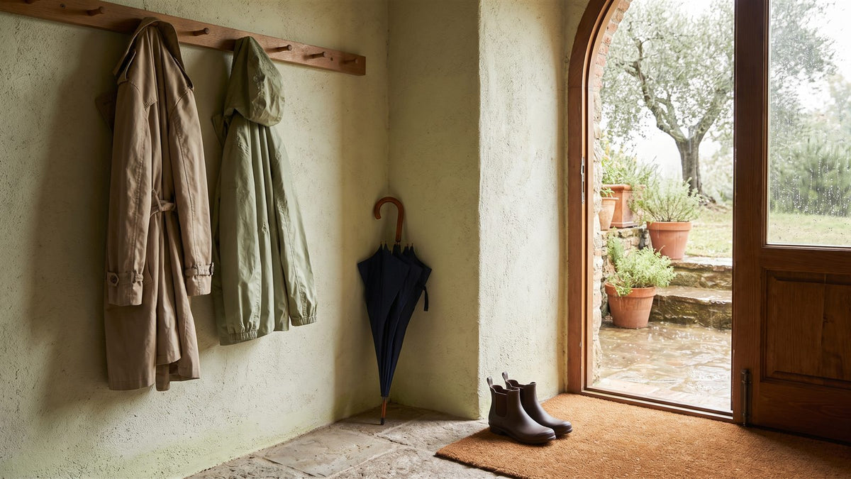 Beige trench coat and green rain jacket hanging by an open door in the rain