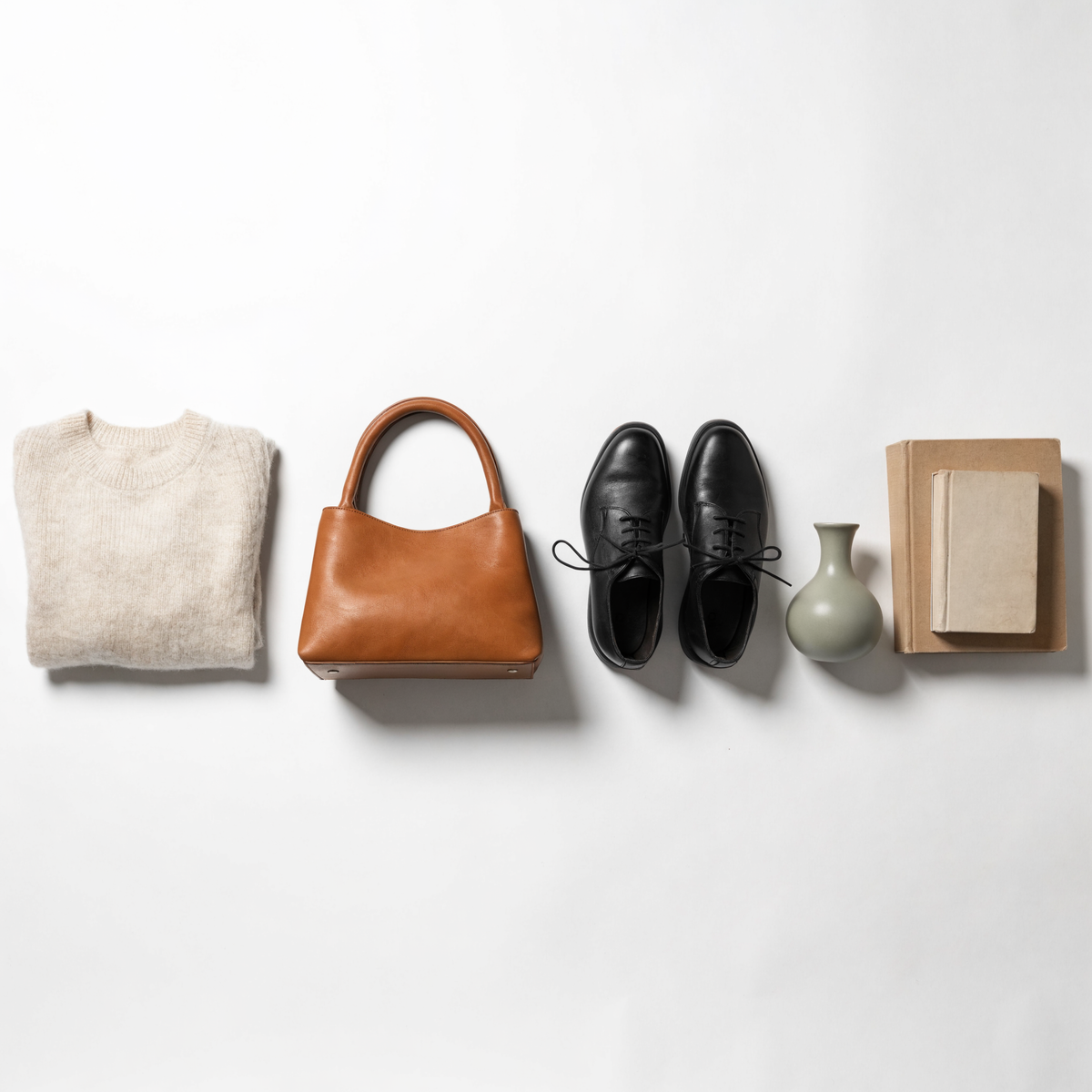 A knit sweater, leather handbag, black shoes, ceramic vase and books laid out on white